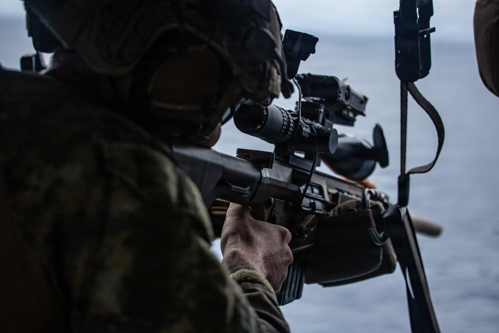 A military service member in camouflage uniform and tactical helmet is aiming a rifle equipped with an optical scope toward an open body of water. The image appears to be taken from an elevated or maritime platform, with a second armed individual partially visible on the right. The scene suggests a maritime security or training exercise.