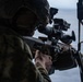 A military service member in camouflage uniform and tactical helmet is aiming a rifle equipped with an optical scope toward an open body of water. The image appears to be taken from an elevated or maritime platform, with a second armed individual partially visible on the right. The scene suggests a maritime security or training exercise.