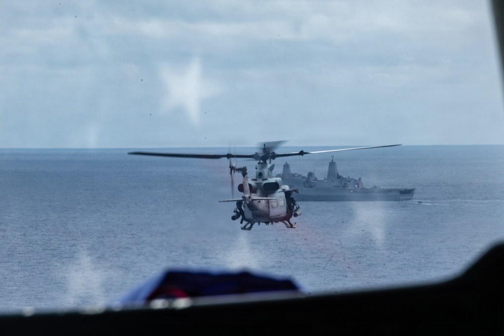 The image is taken from inside an aircraft looking out through a window at a military helicopter hovering over open ocean. A large naval warship, appearing to be an amphibious assault or transport vessel, is visible in the background. The scene suggests a maritime military operation or exercise in progress.