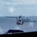 The image is taken from inside an aircraft looking out through a window at a military helicopter hovering over open ocean. A large naval warship, appearing to be an amphibious assault or transport vessel, is visible in the background. The scene suggests a maritime military operation or exercise in progress.