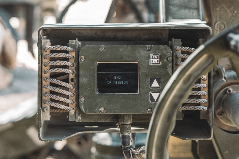 A ruggedized military-grade electronic display unit mounted on what appears to be a vehicle or equipment platform. The small OLED or LCD screen displays the text 'END OF MISSION' in white characters on a black background. The unit features vibration-isolation wire rope mounts on both sides and navigation buttons, consistent with field-use military hardware.