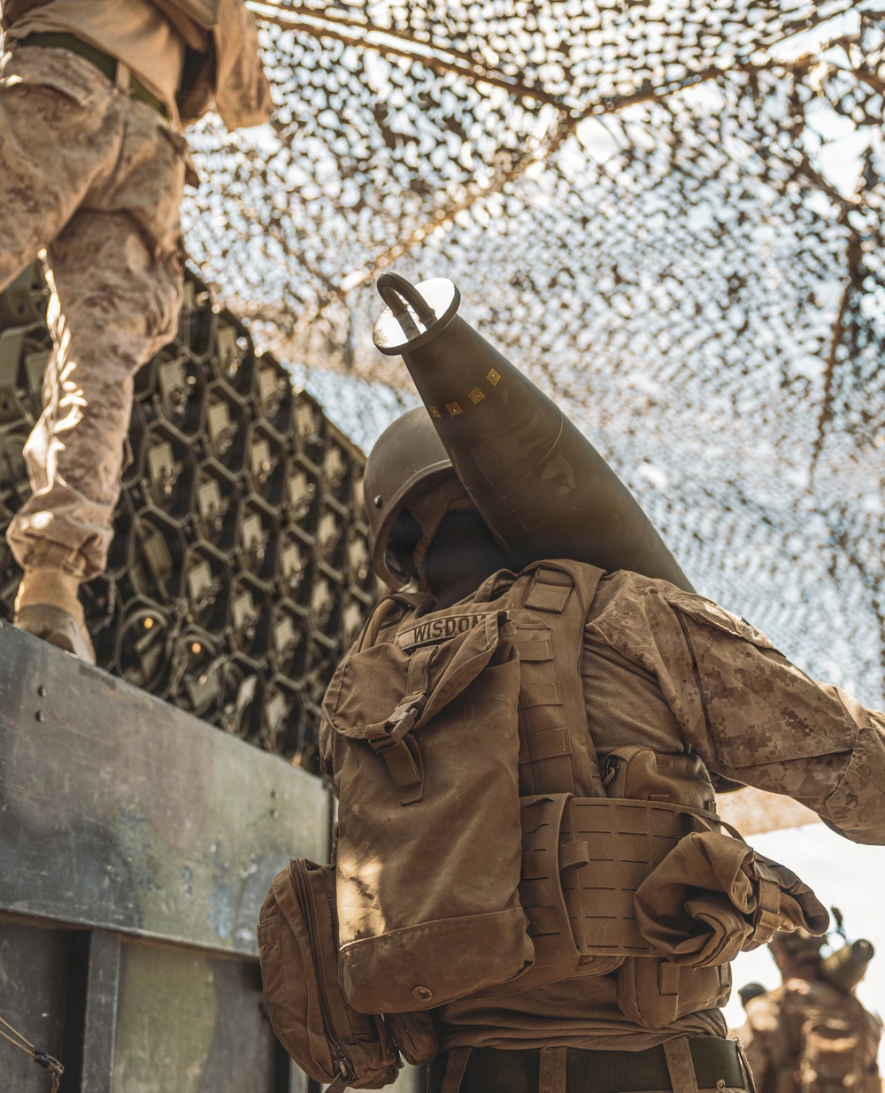 A military service member wearing desert camouflage (MARPAT pattern) and tactical vest with the name tape 'WISDOM' is handling a large artillery or mortar round, holding it upright near their head. The scene is set beneath a camouflage netting canopy, suggesting a field position or forward operating base. A second service member in similar uniform is partially visible in the upper left.