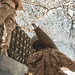 A military service member wearing desert camouflage (MARPAT pattern) and tactical vest with the name tape 'WISDOM' is handling a large artillery or mortar round, holding it upright near their head. The scene is set beneath a camouflage netting canopy, suggesting a field position or forward operating base. A second service member in similar uniform is partially visible in the upper left.