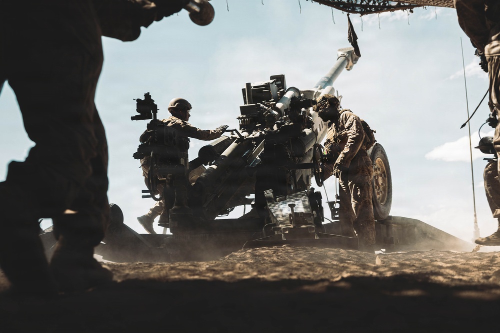Military personnel are operating a towed artillery piece, likely an M777 howitzer or similar 155mm field gun, in an arid outdoor environment. Multiple soldiers in full combat gear and helmets are actively working around the weapon system. The image is taken from a low angle with strong backlighting, creating dramatic silhouettes against a partly cloudy sky.