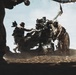 Military personnel are operating a towed artillery piece, likely an M777 howitzer or similar 155mm field gun, in an arid outdoor environment. Multiple soldiers in full combat gear and helmets are actively working around the weapon system. The image is taken from a low angle with strong backlighting, creating dramatic silhouettes against a partly cloudy sky.