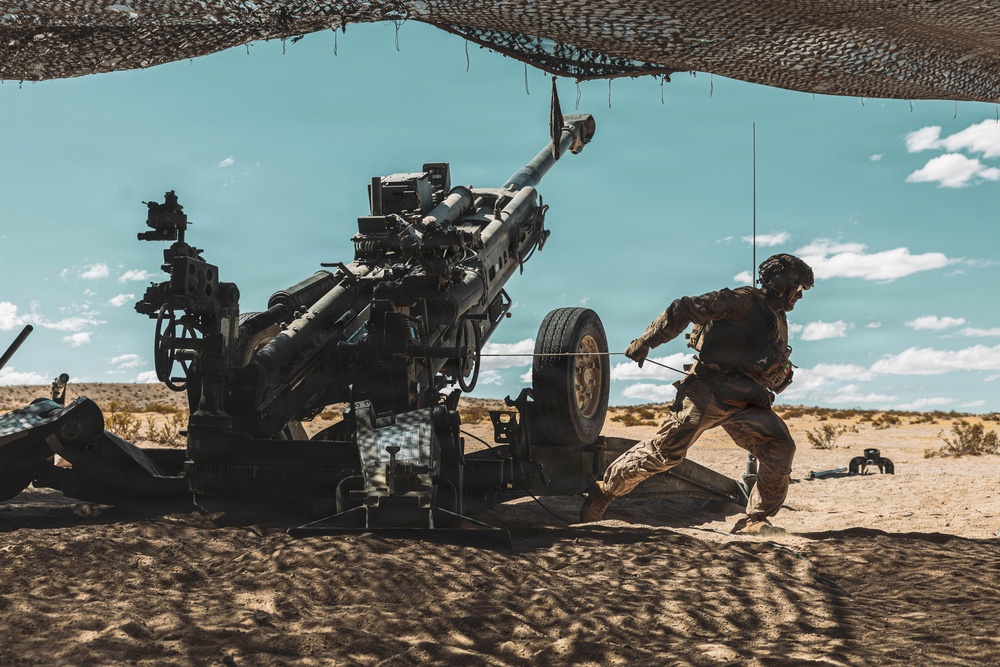 A soldier in desert camouflage uniform is operating a large artillery piece, appearing to pull a lanyard to fire the weapon. The scene takes place in an arid desert environment under a camouflage netting canopy. The setting and equipment suggest a military training exercise or live-fire operation.
