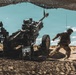A soldier in desert camouflage uniform is operating a large artillery piece, appearing to pull a lanyard to fire the weapon. The scene takes place in an arid desert environment under a camouflage netting canopy. The setting and equipment suggest a military training exercise or live-fire operation.