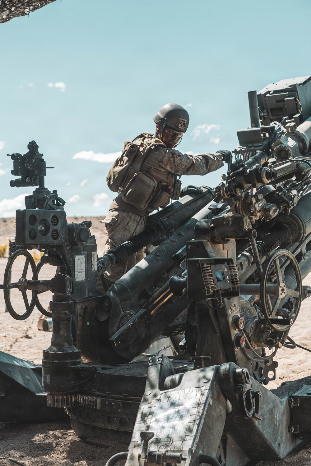 A military service member in desert camouflage uniform and combat helmet is operating or maintaining a large artillery piece in an arid, desert environment. The soldier appears to be working on the breech or upper mechanism of what appears to be an M777 lightweight howitzer. The setting suggests a training exercise or field operation under clear, sunny skies.