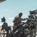 A military service member in desert camouflage uniform and combat helmet is operating or maintaining a large artillery piece in an arid, desert environment. The soldier appears to be working on the breech or upper mechanism of what appears to be an M777 lightweight howitzer. The setting suggests a training exercise or field operation under clear, sunny skies.