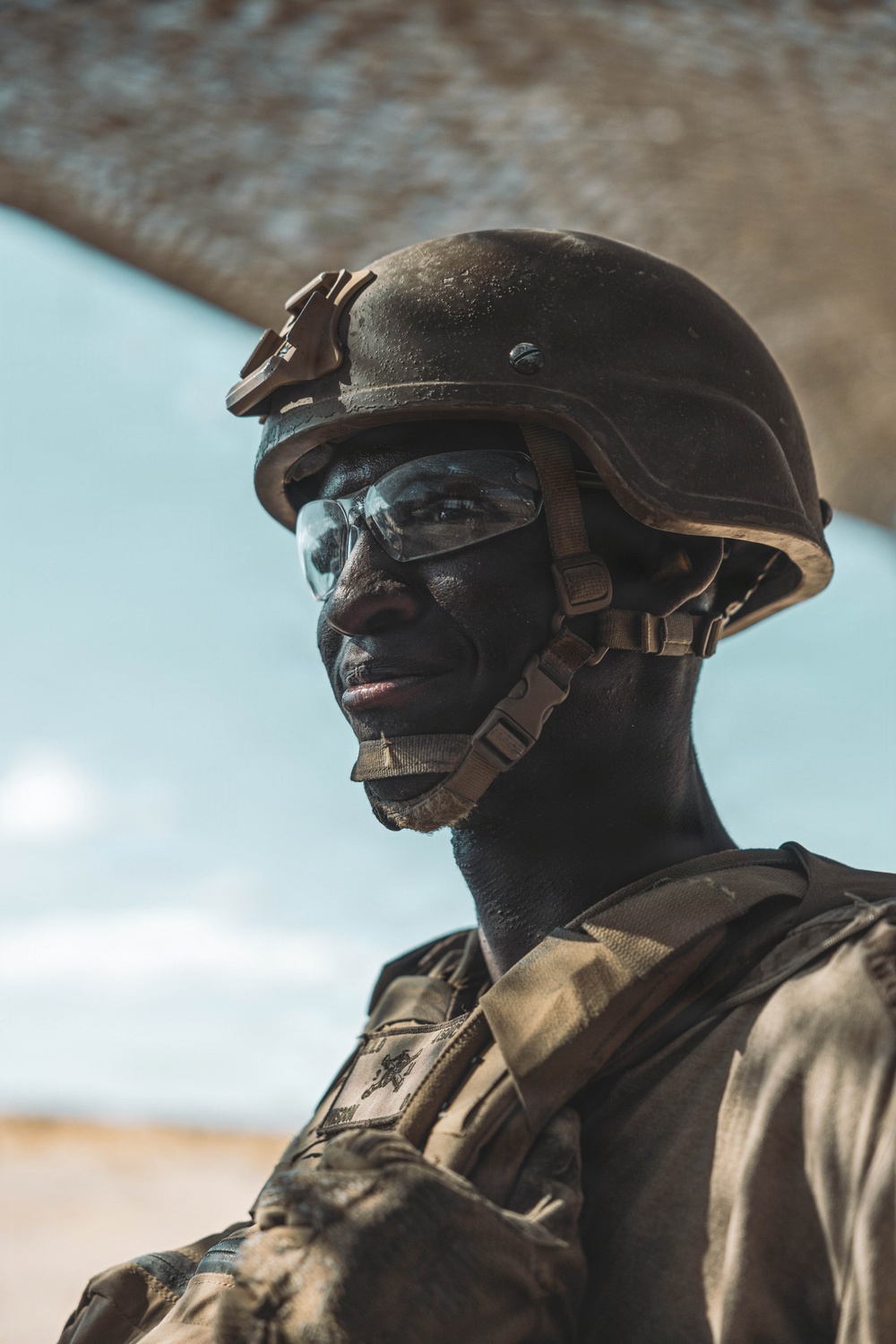 A close-up portrait of a military soldier wearing full combat gear including a ballistic helmet with NVG mount, clear protective eyewear, and tactical vest with a unit patch visible on the chest. The soldier appears to be in an arid or desert environment, with sandy terrain and a blue sky visible in the background. The image is a high-quality, dramatic portrait with strong contrast and natural lighting.