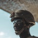 A close-up portrait of a military soldier wearing full combat gear including a ballistic helmet with NVG mount, clear protective eyewear, and tactical vest with a unit patch visible on the chest. The soldier appears to be in an arid or desert environment, with sandy terrain and a blue sky visible in the background. The image is a high-quality, dramatic portrait with strong contrast and natural lighting.