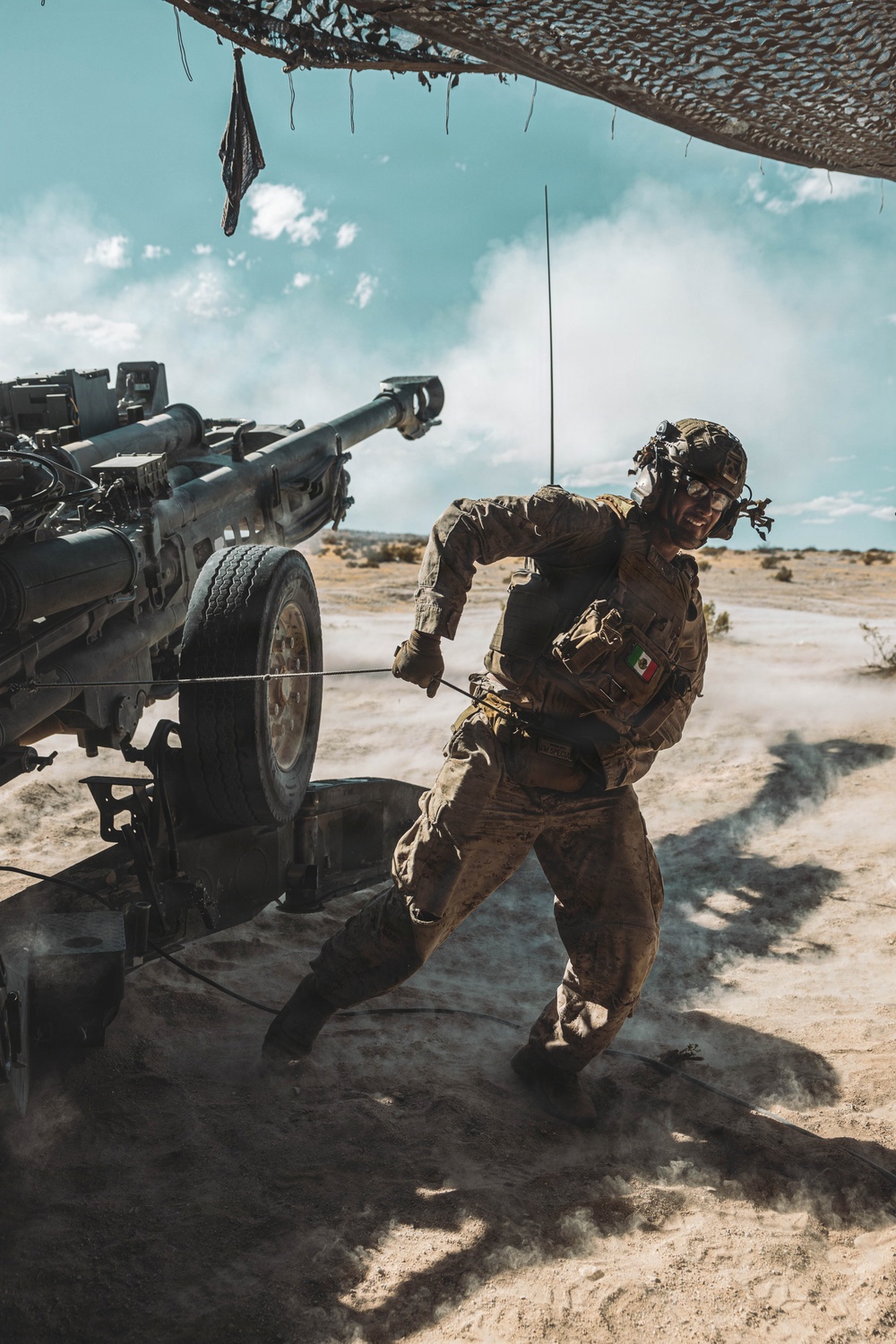 A soldier wearing camouflage uniform and tactical gear is pulling a lanyard cord on what appears to be an M198 or similar towed howitzer artillery piece in a desert environment. The soldier has a Mexican flag patch visible on their left arm, indicating affiliation with the Mexican military. Smoke or dust is visible around the base of the artillery piece, suggesting a recent or imminent firing action.