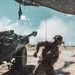 A soldier wearing camouflage uniform and tactical gear is pulling a lanyard cord on what appears to be an M198 or similar towed howitzer artillery piece in a desert environment. The soldier has a Mexican flag patch visible on their left arm, indicating affiliation with the Mexican military. Smoke or dust is visible around the base of the artillery piece, suggesting a recent or imminent firing action.