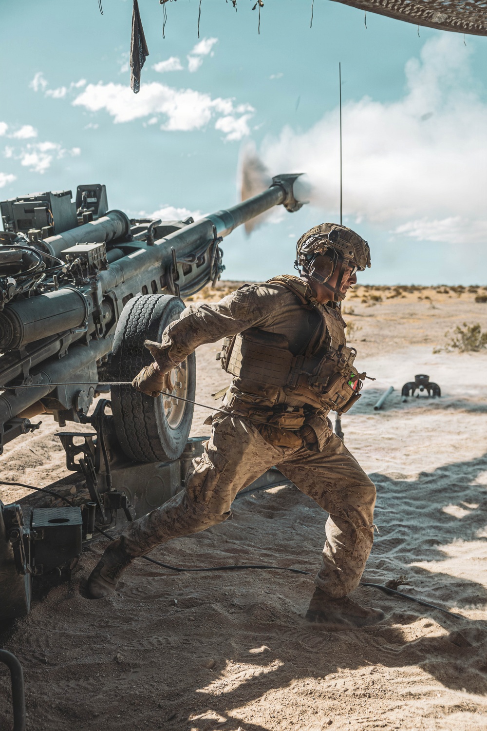 A soldier in desert camouflage uniform is captured mid-action firing a large artillery piece, likely an M777 howitzer, in an arid desert environment. The image captures the moment of discharge with visible muzzle blast smoke at the barrel tip. The soldier is pulling a lanyard to fire the weapon while wearing full combat gear including helmet, plate carrier, and tactical equipment.