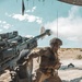 A soldier in desert camouflage uniform is captured mid-action firing a large artillery piece, likely an M777 howitzer, in an arid desert environment. The image captures the moment of discharge with visible muzzle blast smoke at the barrel tip. The soldier is pulling a lanyard to fire the weapon while wearing full combat gear including helmet, plate carrier, and tactical equipment.