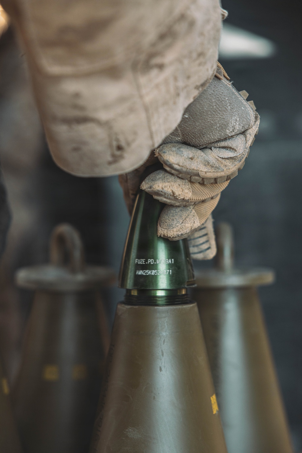 A gloved hand wearing tan/desert-colored tactical gloves is handling a fuze being installed onto an artillery or mortar round. The fuze is marked with visible text identifying it as a point-detonating fuze. The image appears to be a military ordnance handling photograph.