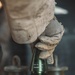 A gloved hand wearing tan/desert-colored tactical gloves is handling a fuze being installed onto an artillery or mortar round. The fuze is marked with visible text identifying it as a point-detonating fuze. The image appears to be a military ordnance handling photograph.