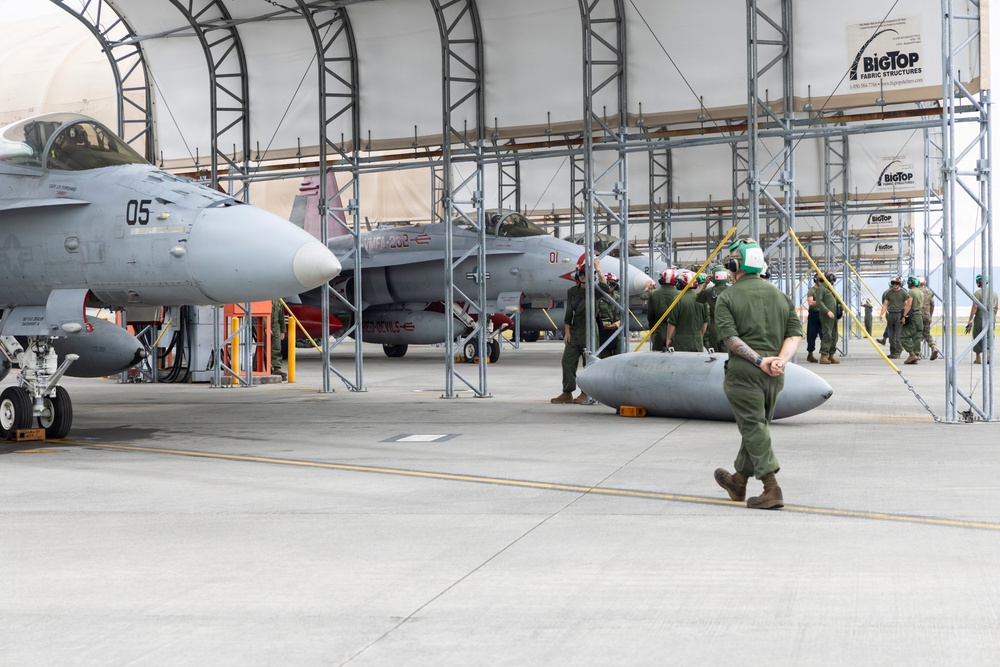 Military personnel are conducting aircraft maintenance and ordnance handling operations inside a fabric hangar structure branded 'BigTop Fabric Structures.' At least two gray military jet aircraft are visible, with ground crews working around them. A munition or external fuel tank is visible on the ground near a walking crew member wearing a green helmet.