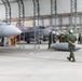 Military personnel are conducting aircraft maintenance and ordnance handling operations inside a fabric hangar structure branded 'BigTop Fabric Structures.' At least two gray military jet aircraft are visible, with ground crews working around them. A munition or external fuel tank is visible on the ground near a walking crew member wearing a green helmet.