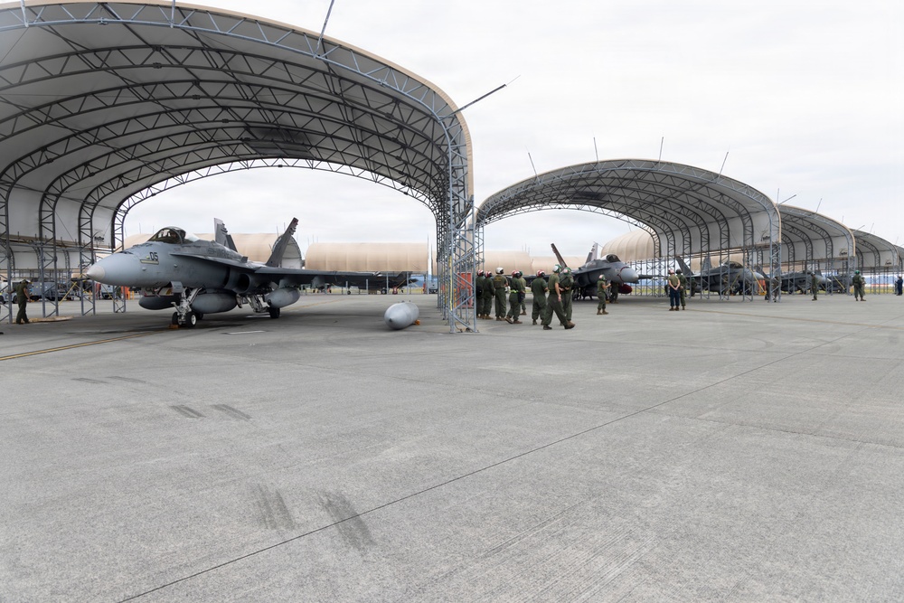 A military airbase tarmac showing multiple jet aircraft parked under large open-sided arched hangar structures. Personnel in flight suits are gathered between the hangars, likely conducting a briefing or post-flight debrief. The scene depicts routine flight operations at what appears to be a well-equipped military air installation.