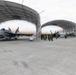 A military airbase tarmac showing multiple jet aircraft parked under large open-sided arched hangar structures. Personnel in flight suits are gathered between the hangars, likely conducting a briefing or post-flight debrief. The scene depicts routine flight operations at what appears to be a well-equipped military air installation.