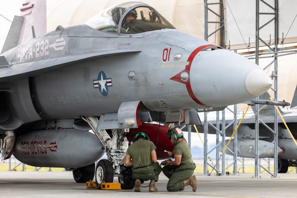A U.S. Marine Corps F/A-18 Hornet fighter jet assigned to VMFA-232 'Red Devils' squadron is shown on a flight line apron. Two ground crew personnel in olive drab uniforms and green helmets are crouched beneath the aircraft conducting a pre-flight or post-flight inspection, while a pilot is visible seated in the cockpit. The aircraft bears modex number '01' and distinctive red devil markings on the nose and external fuel tank.