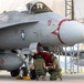A U.S. Marine Corps F/A-18 Hornet fighter jet assigned to VMFA-232 'Red Devils' squadron is shown on a flight line apron. Two ground crew personnel in olive drab uniforms and green helmets are crouched beneath the aircraft conducting a pre-flight or post-flight inspection, while a pilot is visible seated in the cockpit. The aircraft bears modex number '01' and distinctive red devil markings on the nose and external fuel tank.