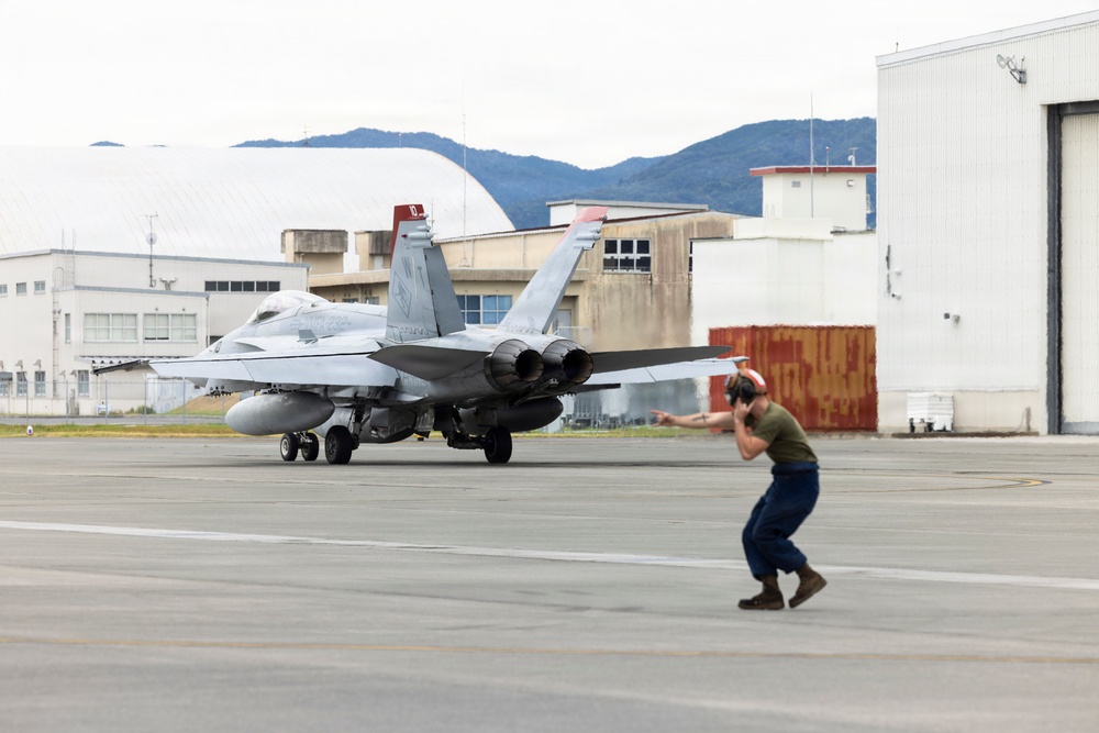 A military jet aircraft, appearing to be an F/A-18 Hornet or Super Hornet, is taxiing on a flight line at what appears to be a military air base. A ground crew member wearing hearing protection is directing the aircraft using hand signals. The background shows military hangars, hardened aircraft shelters, and mountainous terrain suggesting a location in East Asia, possibly Japan.