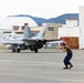 A military jet aircraft, appearing to be an F/A-18 Hornet or Super Hornet, is taxiing on a flight line at what appears to be a military air base. A ground crew member wearing hearing protection is directing the aircraft using hand signals. The background shows military hangars, hardened aircraft shelters, and mountainous terrain suggesting a location in East Asia, possibly Japan.