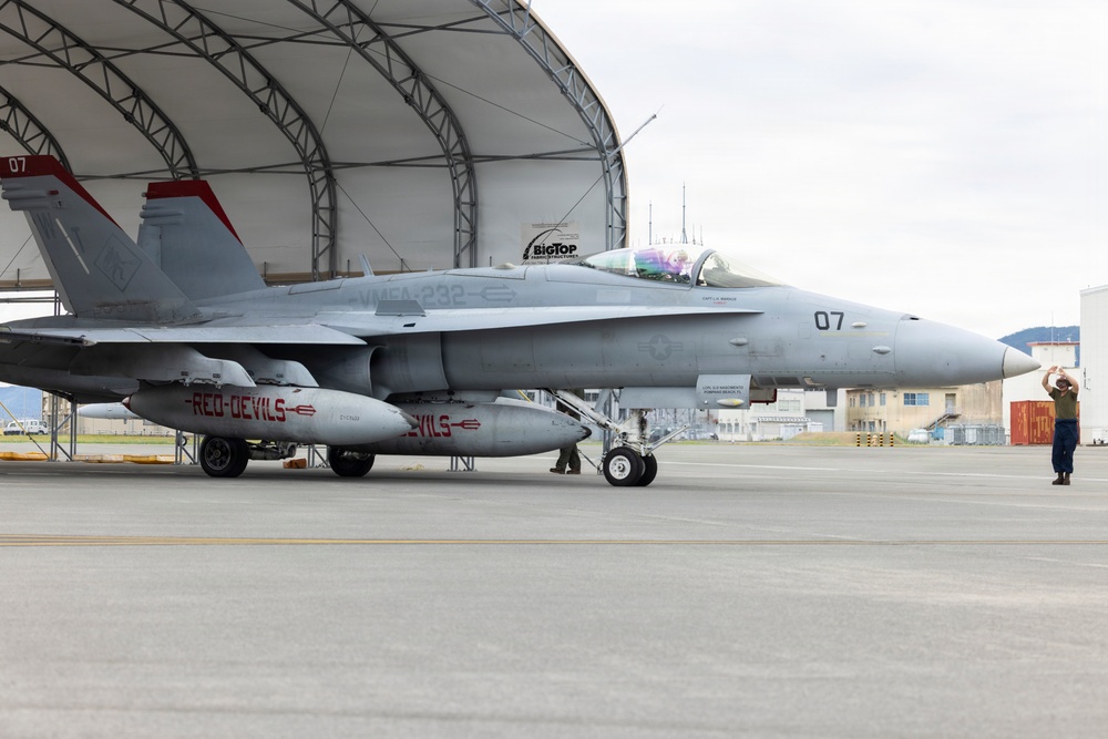 A U.S. Marine Corps F/A-18 Hornet fighter jet assigned to VMFA-232 'Red Devils' squadron, marked with tail number 07, is positioned on a flight line apron in front of a BigTop fabric hangar structure. The aircraft displays 'RED DEVILS' markings on its external fuel/drop tanks and 'VMFA-232' on the fuselage. A ground crew member is visible to the right, appearing to signal or direct the aircraft.
