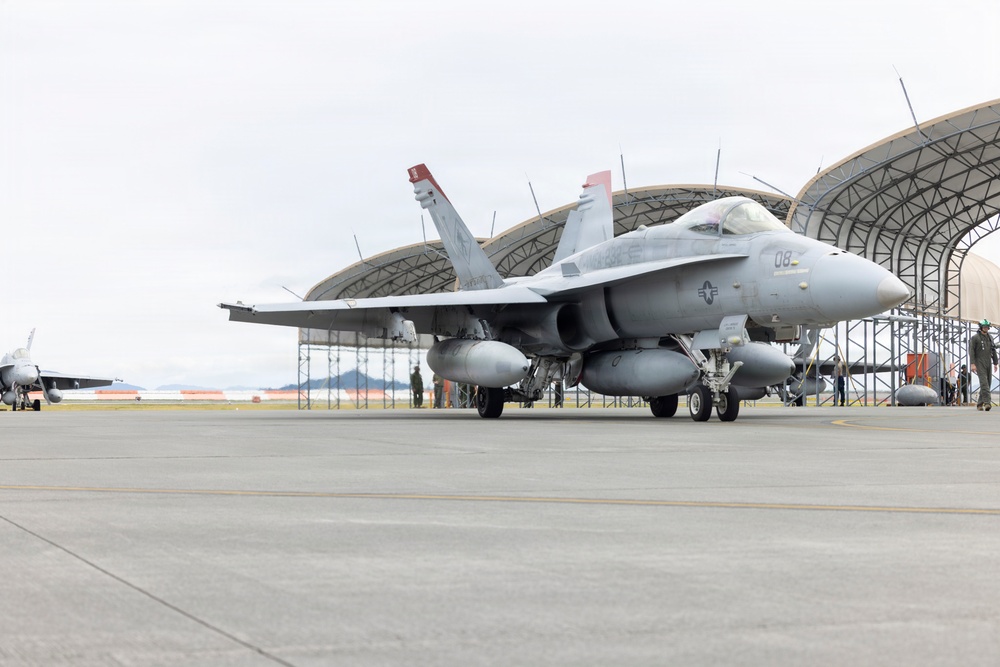 A U.S. military F/A-18 Hornet or Super Hornet jet fighter is parked on a tarmac apron in front of large arched aircraft hangars. The aircraft bears the number '08' on its fuselage and displays U.S. Navy or Marine Corps markings, with external fuel tanks and ordnance pylons visible. Ground crew personnel are visible in the background near the hangars.