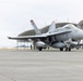 A U.S. military F/A-18 Hornet or Super Hornet jet fighter is parked on a tarmac apron in front of large arched aircraft hangars. The aircraft bears the number '08' on its fuselage and displays U.S. Navy or Marine Corps markings, with external fuel tanks and ordnance pylons visible. Ground crew personnel are visible in the background near the hangars.