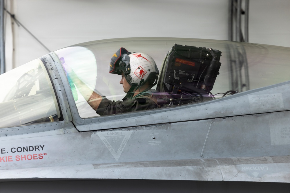 A military pilot is seated in the cockpit of a fighter jet, likely an F-16 or similar aircraft, inside or near a hangar. The pilot is wearing a distinctive white flight helmet with red devil/trident markings and olive drab flight suit. The aircraft fuselage displays the name 'E. CONDRY' and callsign 'SKIE SHOES' in red lettering.