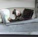 A military pilot is seated in the cockpit of a fighter jet, likely an F-16 or similar aircraft, inside or near a hangar. The pilot is wearing a distinctive white flight helmet with red devil/trident markings and olive drab flight suit. The aircraft fuselage displays the name 'E. CONDRY' and callsign 'SKIE SHOES' in red lettering.