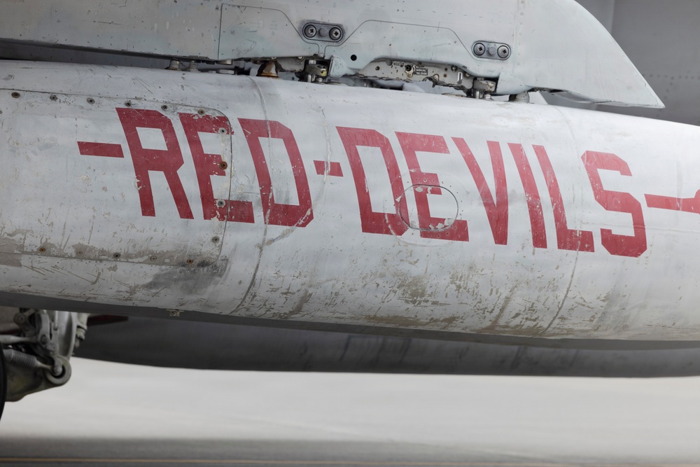 Close-up photograph of a military aircraft fuselage painted white/grey with the text '-RED·DEVILS-' in large red letters. The aircraft shows significant wear, including scratches, paint chipping, corrosion stains, and exposed rivets. Landing gear components are partially visible at the lower left of the frame.