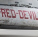Close-up photograph of a military aircraft fuselage painted white/grey with the text '-RED·DEVILS-' in large red letters. The aircraft shows significant wear, including scratches, paint chipping, corrosion stains, and exposed rivets. Landing gear components are partially visible at the lower left of the frame.