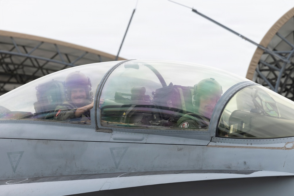 Two military pilots are seated in the cockpit of a two-seat fighter jet, likely an F-16D or similar tandem-seat variant. The front pilot is smiling and appears to be waving or gesturing. The aircraft is parked on a flight line near what appear to be hardened aircraft shelters (HAS) in the background.