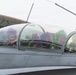 Two military pilots are seated in the cockpit of a two-seat fighter jet, likely an F-16D or similar tandem-seat variant. The front pilot is smiling and appears to be waving or gesturing. The aircraft is parked on a flight line near what appear to be hardened aircraft shelters (HAS) in the background.