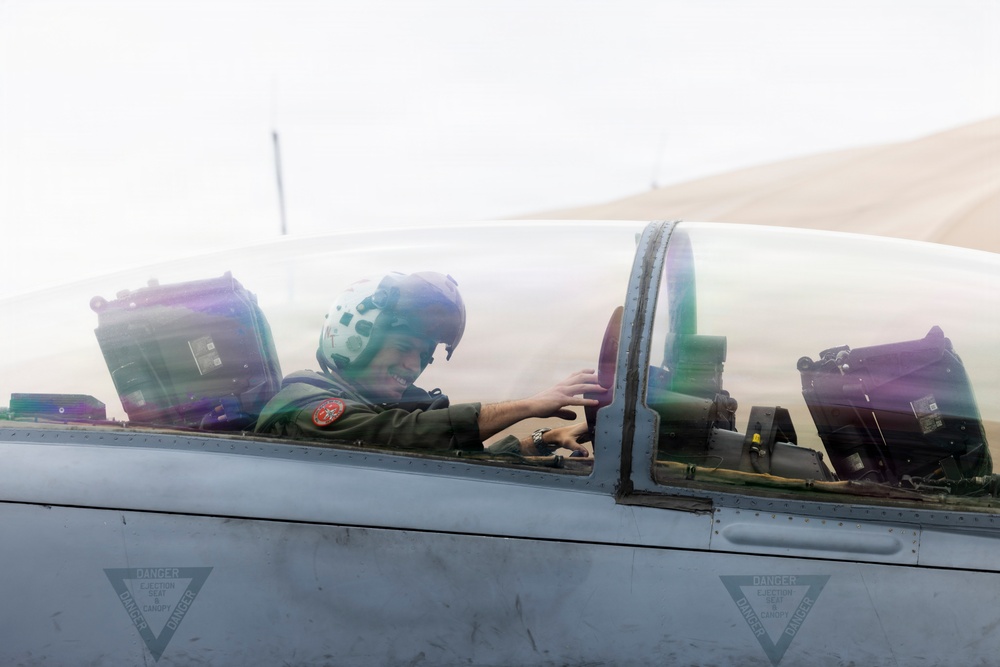 A military pilot is seated in the cockpit of a fighter jet, smiling and appearing relaxed on the ground. The aircraft canopy is partially open, revealing the pilot in a flight suit and helmet. Warning markings for ejection seats are visible on the fuselage.