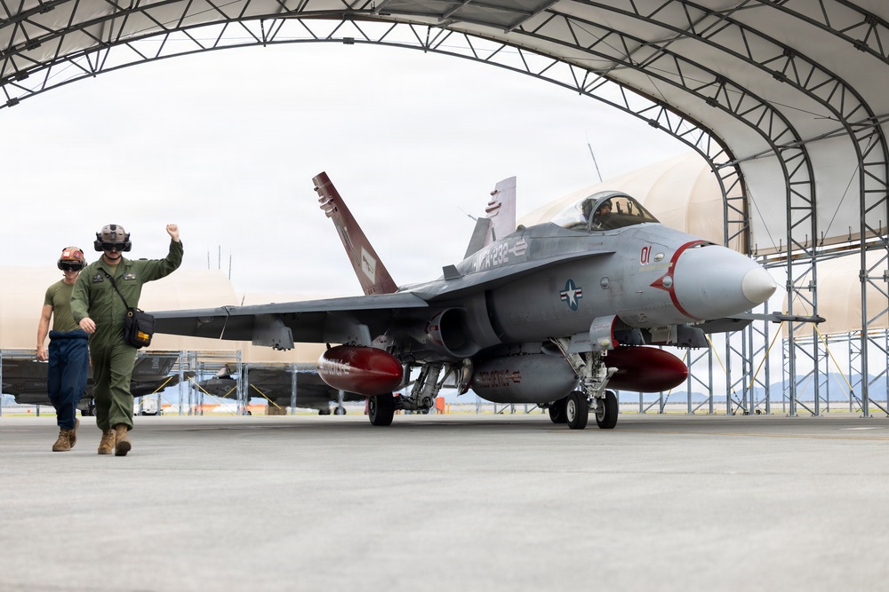 A U.S. Navy F/A-18 Hornet (bureau number visible as '01', tail code 'NR-232') is being taxied out of an arched military hangar. Two personnel in olive green flight suits walk alongside the aircraft, with one raising a fist. The aircraft is equipped with external fuel tanks and appears to be in an operational configuration.