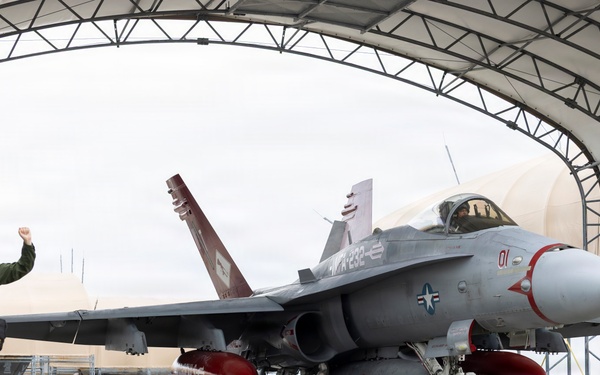 A U.S. Navy F/A-18 Hornet (bureau number visible as '01', tail code 'NR-232') is being taxied out of an arched military hangar. Two personnel in olive green flight suits walk alongside the aircraft, with one raising a fist. The aircraft is equipped with external fuel tanks and appears to be in an operational configuration.