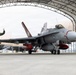 A U.S. Navy F/A-18 Hornet (bureau number visible as '01', tail code 'NR-232') is being taxied out of an arched military hangar. Two personnel in olive green flight suits walk alongside the aircraft, with one raising a fist. The aircraft is equipped with external fuel tanks and appears to be in an operational configuration.