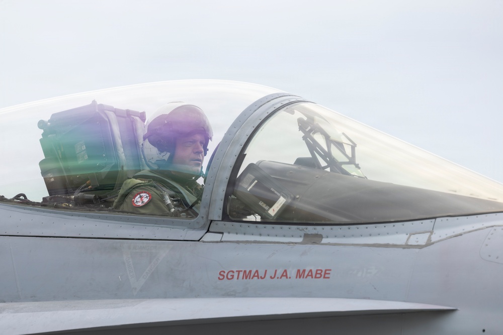 A military pilot is seated in the cockpit of a fighter jet, wearing a flight helmet and olive green flight suit. The aircraft fuselage displays the text 'SGTMAJ J.A. MABE' in red lettering, indicating the crew chief or dedicated crew member's name. The canopy is closed and the pilot appears to be preparing for or returning from a flight.