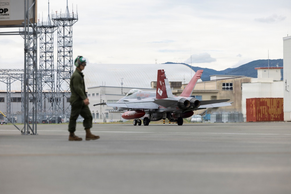 A military jet aircraft, appearing to be an F/A-18 Hornet with distinctive red and white tail markings, is taxiing on a flight line at a military airbase. A flight crew member in olive green flight suit and helmet walks in the foreground. The setting appears to be an overseas installation, likely in Japan based on the architectural style of surrounding buildings and mountainous terrain.