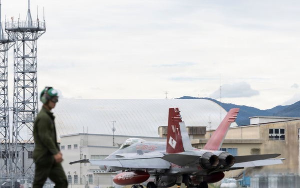 A military jet aircraft, appearing to be an F/A-18 Hornet with distinctive red and white tail markings, is taxiing on a flight line at a military airbase. A flight crew member in olive green flight suit and helmet walks in the foreground. The setting appears to be an overseas installation, likely in Japan based on the architectural style of surrounding buildings and mountainous terrain.
