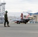 A military jet aircraft, appearing to be an F/A-18 Hornet with distinctive red and white tail markings, is taxiing on a flight line at a military airbase. A flight crew member in olive green flight suit and helmet walks in the foreground. The setting appears to be an overseas installation, likely in Japan based on the architectural style of surrounding buildings and mountainous terrain.