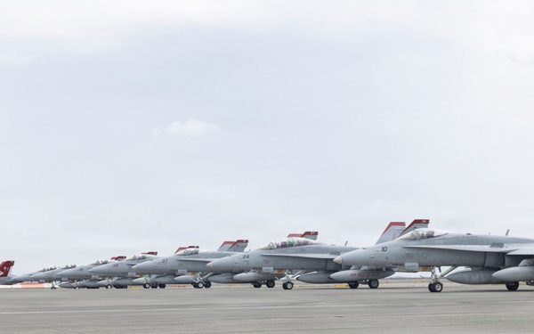 A line of military jet aircraft, appearing to be F/A-18 Hornets or Super Hornets, are parked in formation on a concrete tarmac under an overcast sky. The aircraft are painted in standard gray livery with distinctive red-tipped tail fins and safety covers. The scene appears to be at a naval or military air station, with mountains faintly visible in the background.
