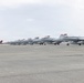 A line of military jet aircraft, appearing to be F/A-18 Hornets or Super Hornets, are parked in formation on a concrete tarmac under an overcast sky. The aircraft are painted in standard gray livery with distinctive red-tipped tail fins and safety covers. The scene appears to be at a naval or military air station, with mountains faintly visible in the background.