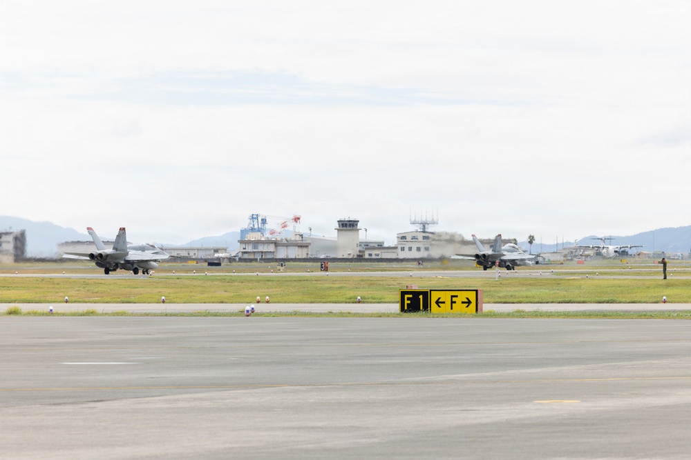 Two military jet aircraft, appearing to be F/A-18 Hornets, are operating on a runway at a military airbase. A yellow taxiway sign reading 'F1' with directional arrows is prominently visible in the foreground. The background shows an air traffic control tower, base infrastructure, and mountainous terrain.