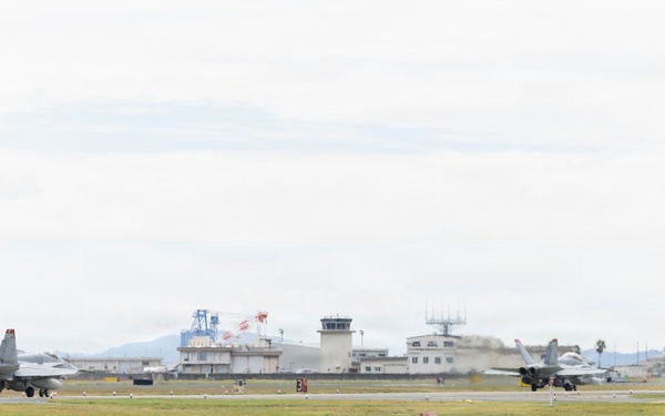 Two military jet aircraft, appearing to be F/A-18 Hornets, are operating on a runway at a military airbase. A yellow taxiway sign reading 'F1' with directional arrows is prominently visible in the foreground. The background shows an air traffic control tower, base infrastructure, and mountainous terrain.